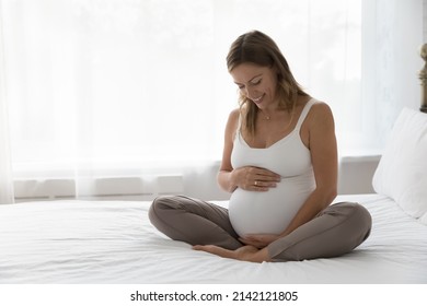 Happy expectant mother sitting on bed, feeling kicks, caressing big belly, smiling. Pregnant woman in white tank top touching bump, speaking to unborn baby. Motherhood, pregnancy concept