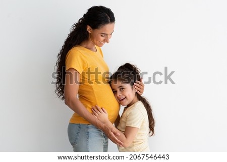Happy Expectation. Adorable Little Preteen Girl Cuddling Mother's Pregnant Belly, Excited Curious Cute Female Child Awaiting For Sibling, Hugging Mommy's Tummy While Bonding Together At Home