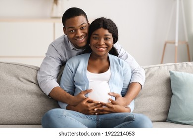 Happy expecting african american couple cuddling while spending time together at home, copy space. Pregnant black couple sitting on couch at living room and hugging woman big belly, smiling at camera