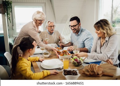 Happy Extended Family Having Lunch Together In Dining Room. Senior Woman Is Serving Food At The Table. 