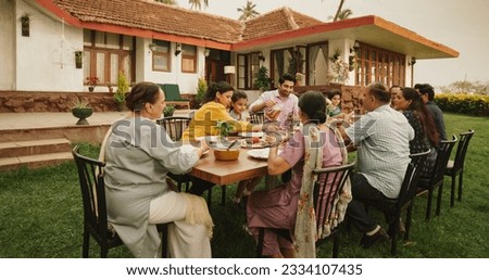 Happy Extended Indian Family Eating Together Outdoors in House Backyard. Grandparents, Parents, and Children Enjoying Each Other's Company Over a Big Table Full of Traditional Delicious Food