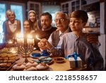 Happy extended Jewish family celebrating Hanukkah while gathering at dining table. Focus is on boy lighting candles in menorah.
