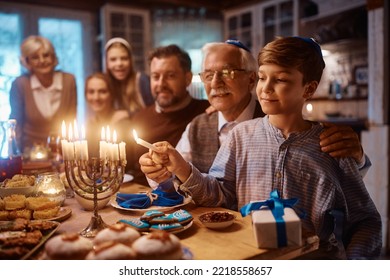 Happy extended Jewish family celebrating Hanukkah while gathering at dining table. Focus is on boy lighting candles in menorah.