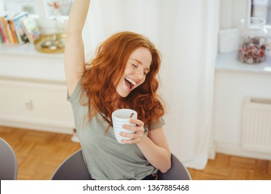 Happy extrovert carefree young woman cheering raising her arm and laughing as she holds a mug of coffee indoors at home