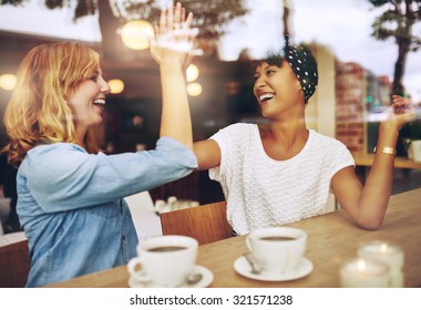Happy exuberant young girl friends giving a high five slapping each others hand in congratulations as they sit together in a cafeteria enjoying a cup of hot coffee, multi ethnic viewed through glass