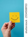 Happy face icon on a blue background. A hand holds a yellow smiley face icon on a bright blue background. This cheerful image conveys positivity and optimism.