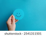 Happy face icon on a blue background. A hand holds a smiley face icon on a bright blue background. This cheerful image conveys positivity and optimism.