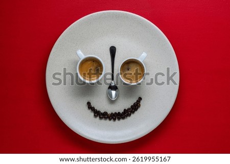 Happy face shape made from two white cups of espresso coffee, teaspoon and coffee beans in plate on red background, close up, top view. Creative composition of funny food