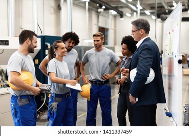 Happy Factory Worker And Her Colleagues Showing Business Reports To Company Leaders While Having A Meeting In Industrial Plant.