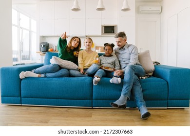 Happy Familly Mother, Father And Two Daughters Sitting On Sofa And Watching Tv At Home. Adoption Concept