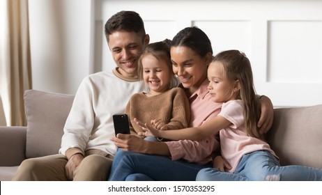 Happy Family Adult Parents With Cute Small Kids Daughters Relax On Sofa Laughing Having Fun Looking At Phone Screen, Mum Dad And Children Watch Funny Video Take Selfie Play Game On Smartphone At Home