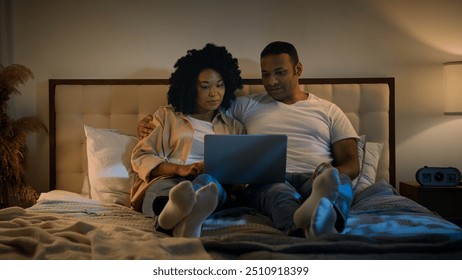 Happy family African American married couple relax at evening at home bedroom on bed man and woman at night browsing laptop using computer talking discuss shopping together online watching movie film - Powered by Shutterstock