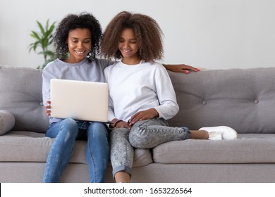 Happy family african american mother and teen daughter looking at laptop sitting on sofa in modern living room having fun making video call, doing online shopping or watching movie using computer