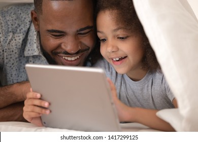 Happy Family African Father And Cute Small Kid Daughter Using Digital Tablet Lying On Bed Under Blanket, Funny Little Child Girl With Dad Having Fun Looking At Computer Covered With Duvet In Bedroom