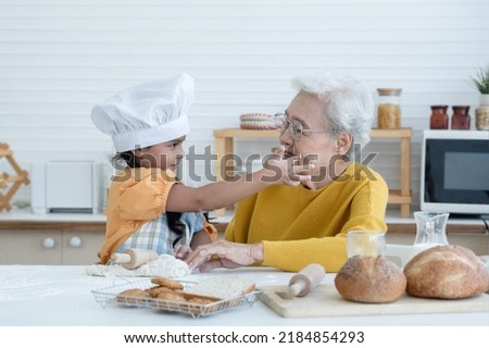 Happy family Asian grandmother and little grandchild spend time together at kitchen, knead dough and bake cookies or bread, niece have fun to tease and put flour on her grandma face at home