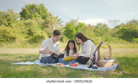 Happy Family Asian With Little Girl Learning Book Together And Have Enjoyed Ourselves Together During Picnicking On A Picnic Cloth In The Green Garden. Family Enjoying Sunny Fall Day In Nature.