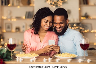 Happy Family. Beautiful Lovely Young African American Couple Using Smartphone, Sitting At Table And Having Dinner. Smiling Woman Sharing Photos With Man Who Hugging Her, Spending Time Together