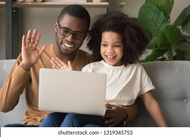 Happy family black dad with kid daughter waving hands making distance video call looking at laptop, african father and child girl looking talking to webcamera chatting online by computer webcam