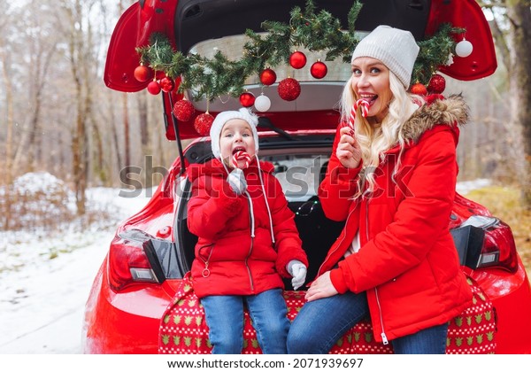 Happy family of\
blonde mom and little girl sit in christmas decorated car in winter\
forest and eat candy\
canes.