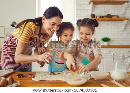 Happy family busy in kitchen. Mother and two helpful little daughters in aprons making cookies on weekend at home. Mom teaches inquisitive children to break egg, prepare dough and use cooking utensils