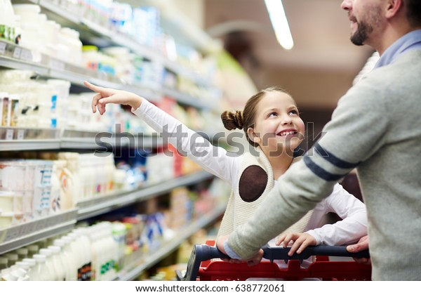 Happy family buying groceries: smiling little\
girl choosing dairy products from fridge in milk aisle while\
shopping in supermarket