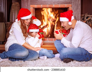 Happy Family By Fireplace, Parents With Baby Girl Wearing Santa Claus Hats, Sitting Relaxed In Ski Resort Chalet And Drinking Hot Tea, Joyful Winter Vacation, People Traveling On Christmas Holidays