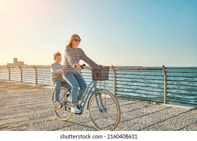 Happy Family, Carefree Mother And Son With Bike Riding On Beach Having Fun, On The Seaside Promenade On A Summer Day, Enjoying Vacation. Togetherness Friendly Concept