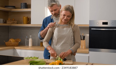 Happy family Caucasian middle-aged adult at kitchen woman cook vegetable salad cut cucumber husband man hugging cuddling embracing wife couple talking discuss diet food affectionate love talk cooking - Powered by Shutterstock