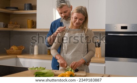 Happy family Caucasian middle-aged adult at kitchen woman cook vegetable salad cut cucumber husband man hugging cuddling embracing wife couple talking discuss diet food affectionate love talk cooking