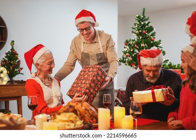 Happy Family Celebrating Christmas Dinner At Home. Father With Gift Box Given To Mother Or Grandmother, Grandfather Wearing Santa Hats.Merry Christmas Or New Year's Eve Concept