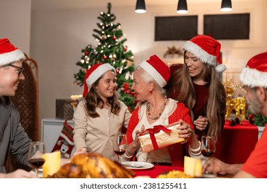 Happy family celebrating Christmas dinner at home. Granddaughter with gift box surprise given to grandmother wearing Santa hats. Merry Christmas or New Year's eve concept. - Powered by Shutterstock