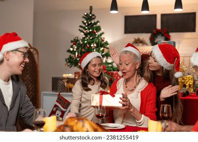 Happy family celebrating Christmas dinner at home. Granddaughter with gift box surprise given to grandmother wearing Santa hats. Merry Christmas or New Year's eve concept. - Powered by Shutterstock