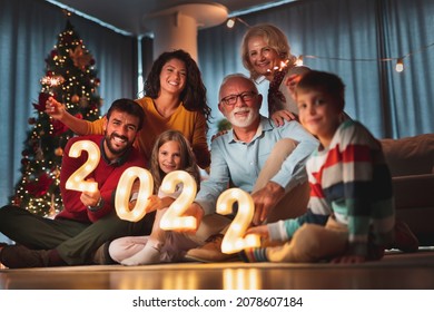 Happy Family Celebrating New Years Eve At Home With Kids, Sitting By The Christmas Tree, Holding Sparklers And Illuminative Numbers 2022 Representing The Upcoming New Year