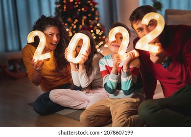 Happy Family Celebrating New Years Eve At Home With Kids, Sitting By The Christmas Tree, Holding  Illuminative Numbers 2022 Representing The Upcoming New Year