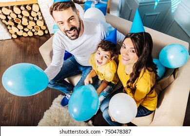 Happy Family. Charming Young Family Sitting On The Sofa Together And Holding Balloons While Wearing Party Hats And Smiling At The Camera