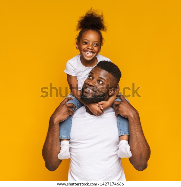 幸せな家族 オレンジのスタジオ背景にかわいい娘を肩に乗せた明るい黒人 の写真素材 今すぐ編集
