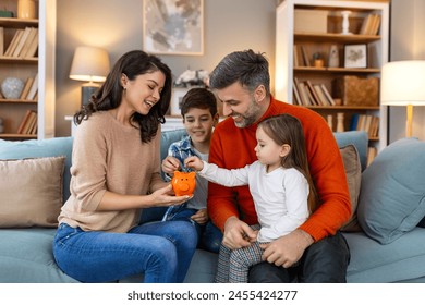 Happy family cheerful mother and father with kids smiling and putting coins into piggy bank while sitting on sofa at home - Powered by Shutterstock