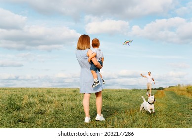 Happy Family With Child And Dog Are Having Fun Outdoors, Mom With Son In Arms Watch As Dad Launches Rainbow Kite Into Blue Sky On Field. Family Vacation, Spending Time Together, Parents With Baby Boy.