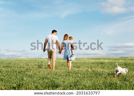 Happy family with child and dog walk in field. Back view of young father holding his wife by hand, mom is carrying her little son, jack russell terrier running on green lawn. Family outdoor recreation