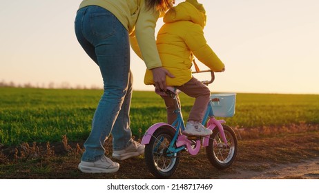 Happy family, child and mom, kid learns to ride a bike in the park at sunset. Mother teaches her daughter, son to ride child's bike on the autumn road. Happy young family, childhood, active lifestyle