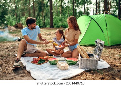 Happy family with a child on a picnic sitting on a blanket near the tent and eating food fried at the stake during the weekend in the forest. Camping, recreation, hiking. - Powered by Shutterstock