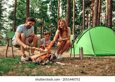 Happy family with a child on a picnic sit by the fire near the tent and grill a barbecue in a pine forest. Camping, recreation, hiking. - Shutterstock ID 2051440751