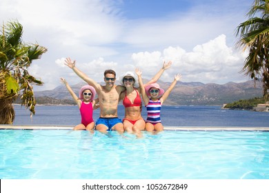 Happy Family With Children Sitting On The Edge Of The Pool In A Tropical Seaside Resort Against The Backdrop Of The Sea And Palms