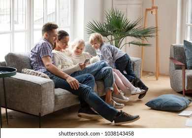 Happy Family With Children Using Mobile Apps Together At Home, Young Couple And Kids Having Fun Playing Game On Smartphone Sitting On Sofa, Parents And Son Daughter Relaxing In Living Room With Phone