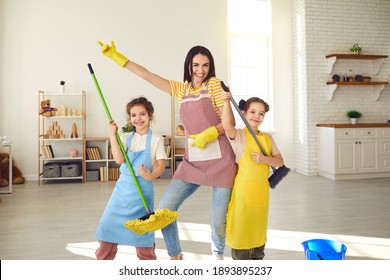 Happy Family Cleaning House And Having Fun. Children Helping Mother Tidy Up The Flat. Smiling Young Mum And Little Daughters Pretend To Play Music Holding Mops Like Guitars While Mopping Floor At Home