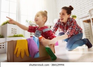 Happy Family Cleans The Room. Mother And Daughter Do The Cleaning In The House. A Young Woman And A Little Child Girl Having Fun And Riding In Cardboard Boxes At Home.