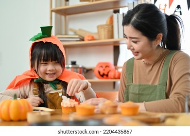 Happy Family Concept. Playful And Cute Young Asian Girl In Halloween Costume Enjoys Making A Halloween Cupcake With Her Mom.