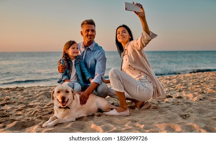 Happy family concept. Young attractive mother, handsome father and their little cute daughter sitting together on the beach with dog and making selfie on a smart phone.