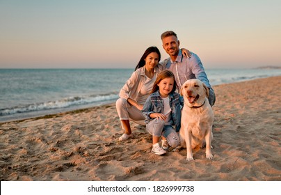 Happy family concept. Young attractive mother, handsome father and their little cute daughter sitting together on the beach with dog.