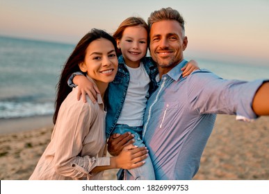 Happy family concept. Young attractive mother, handsome father and their little cute daughter standing together on the beach and making selfie. - Powered by Shutterstock